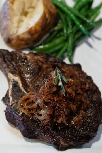 A bone-in ribeye under caramelised onions and a thyme sprig, with a baked potato and green beans behind