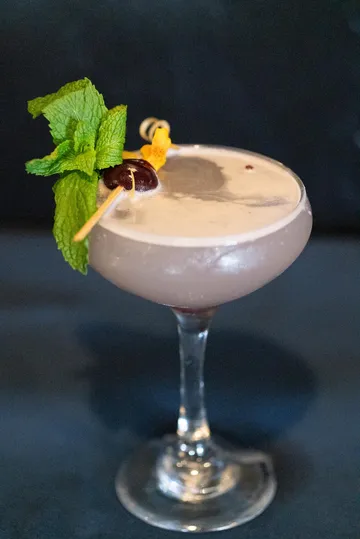 A pale foamy cocktail in a coupe glass, garnished with mint, a dark cherry and an edible flower