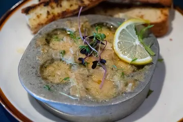 Escargot baked in garlic butter in a metal dish, topped with micro greens and a lemon slice, grilled bread behind
