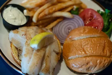 A grilled grouper fillet on a bun with tartar sauce, fries, lettuce, tomato and red onion