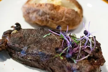 A New York strip steak topped with purple micro greens, a buttered baked potato behind it